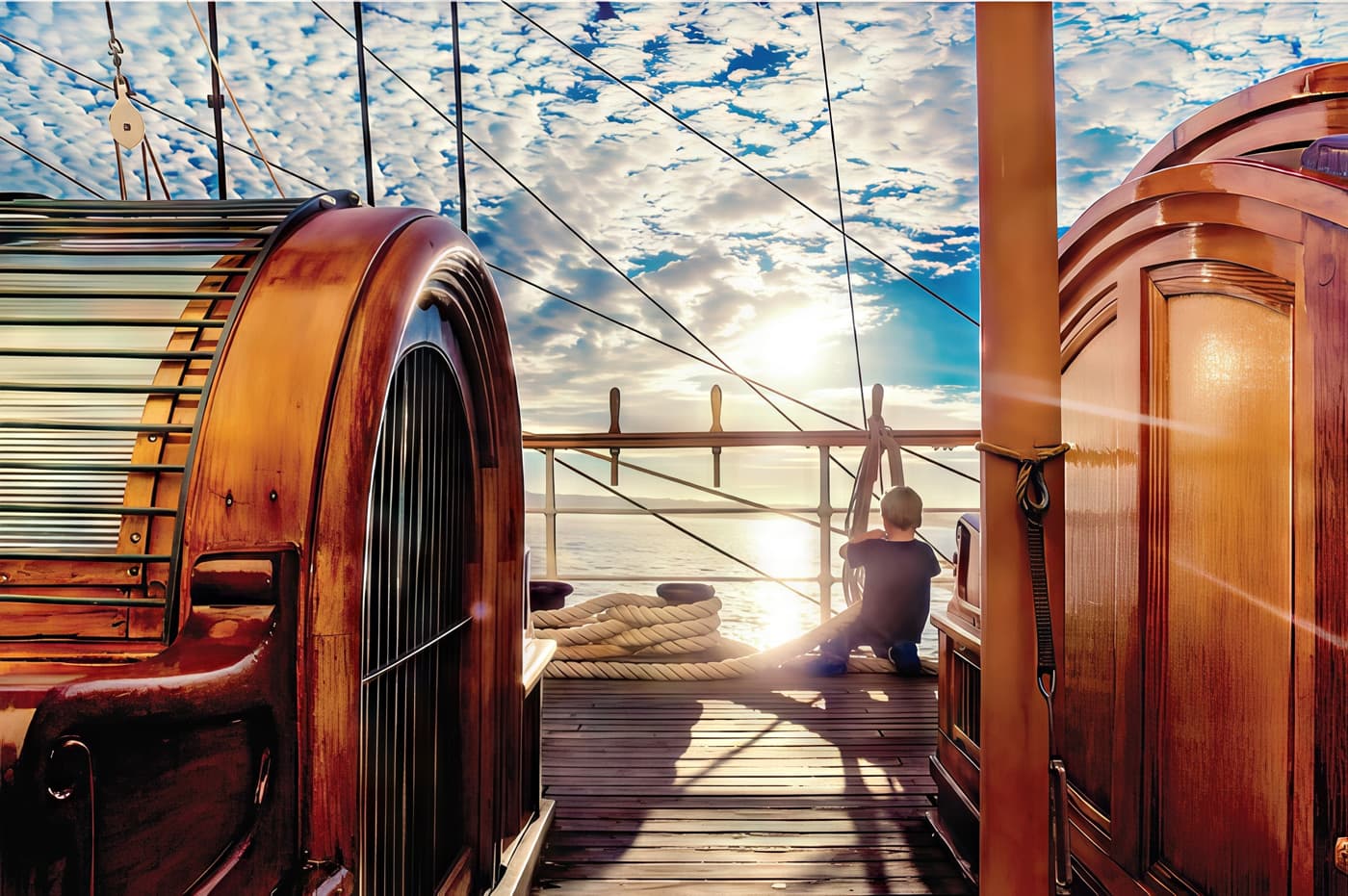 A child sits on the deck of the historic sailing ship Star of India at sunset, surrounded by polished wooden structures and coiled rope, gazing out over the calm ocean under a sky filled with scattered clouds and warm light.